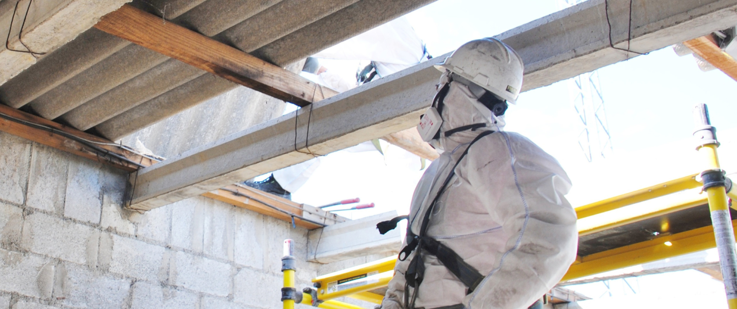 Desmontando tejado de fibrocemento con amianto en Estación Elé Ein Bauarbeiter in Schutzkleidung betrachtet die Decke auf einer Baustelle mit Gerüst und Betonwänden.